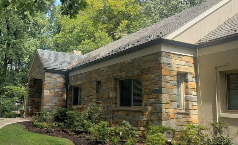 Exterior view of a nursing home building with stone walls, multiple windows, a shingled roof, and surrounding greenery including bushes and trees.