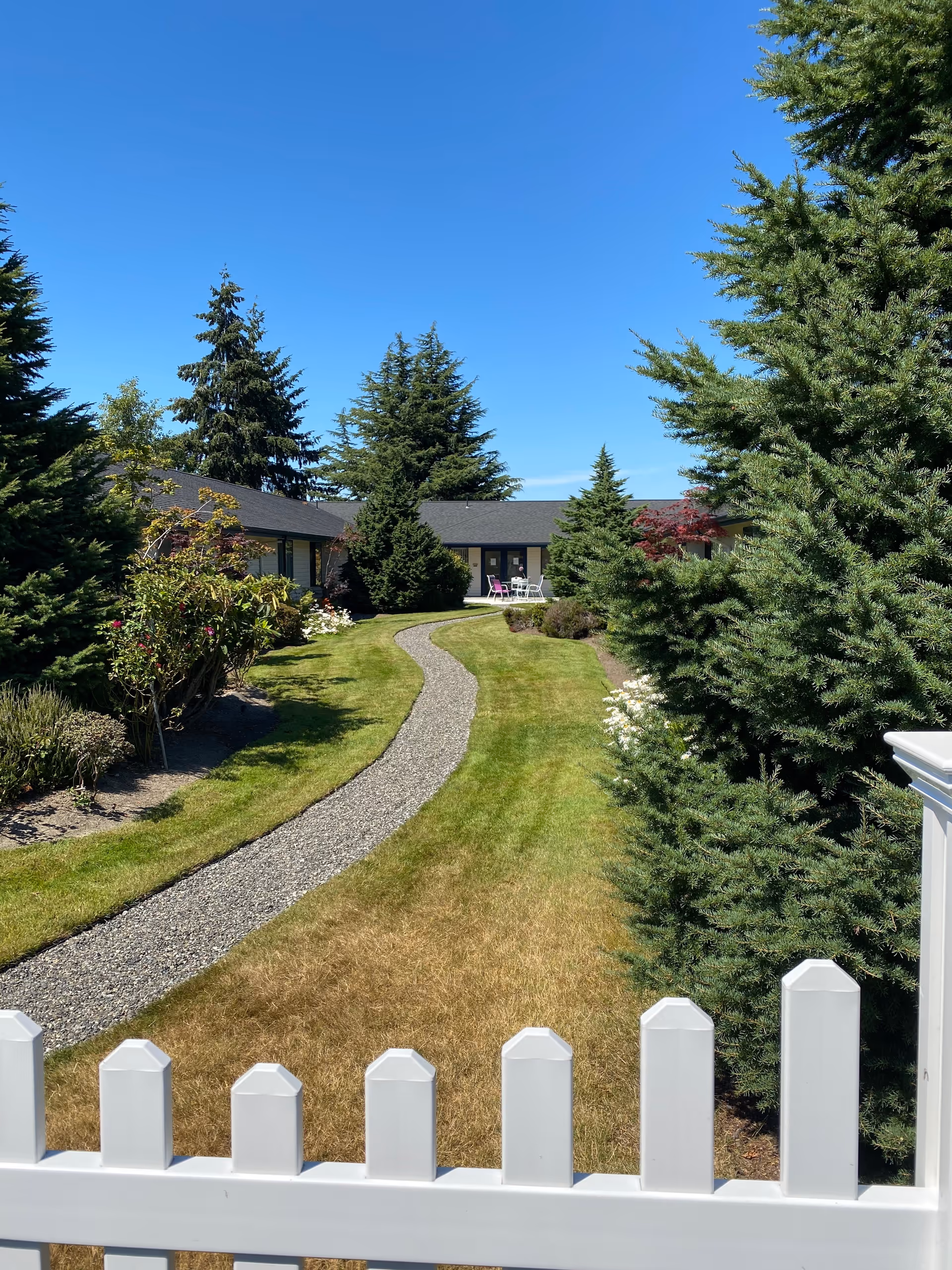 A landscaped outdoor area at Logan Creek Retirement Community featuring a winding gravel path through a green lawn, surrounded by various trees and shrubs, with a white picket fence in the foreground and a building with a patio and chairs in the background under a clear blue sky.
