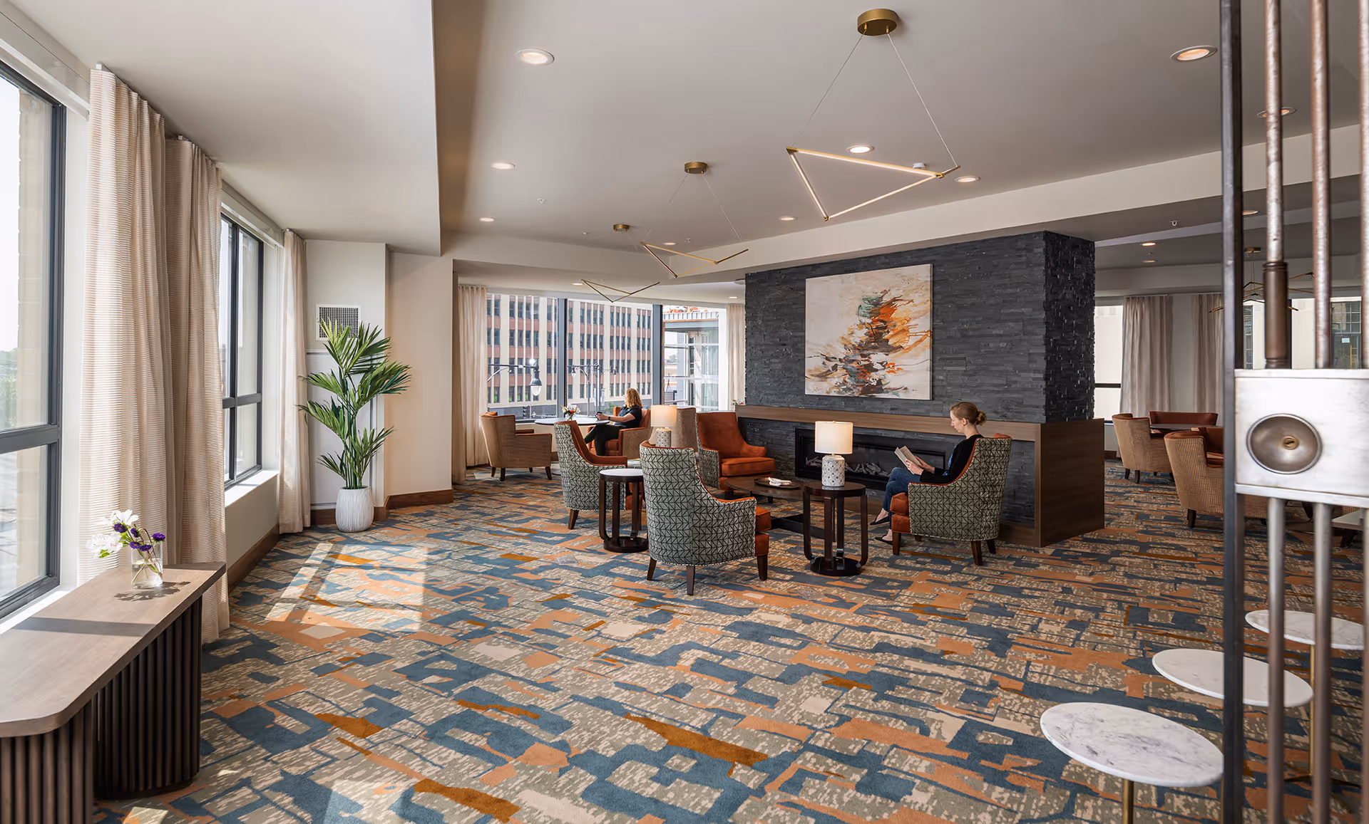 A spacious, well-lit lounge area with large windows letting in natural light. The room features patterned carpet in blue, orange, and beige tones, modern armchairs in green and rust colors arranged around small tables with lamps, and a dark stone fireplace with abstract artwork above it. Two women are seated, one reading a book and the other looking out the window. There are beige curtains, a potted plant, and contemporary geometric pendant lights hanging from the ceiling.