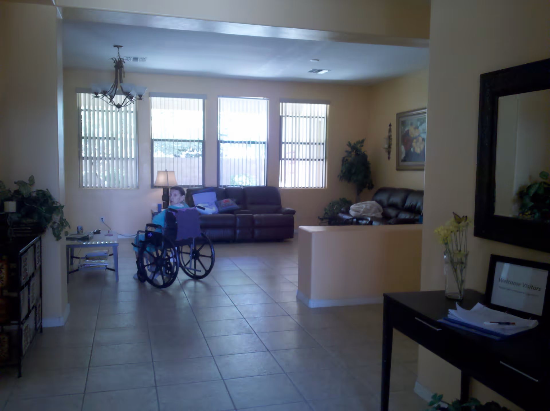 A spacious living room with tiled floors, beige walls, and three large windows with vertical blinds. There are two dark leather sofas, a side table with a lamp, and a woman in a wheelchair facing away from the camera. The room is decorated with plants, a framed painting, and a chandelier hanging from the ceiling.
