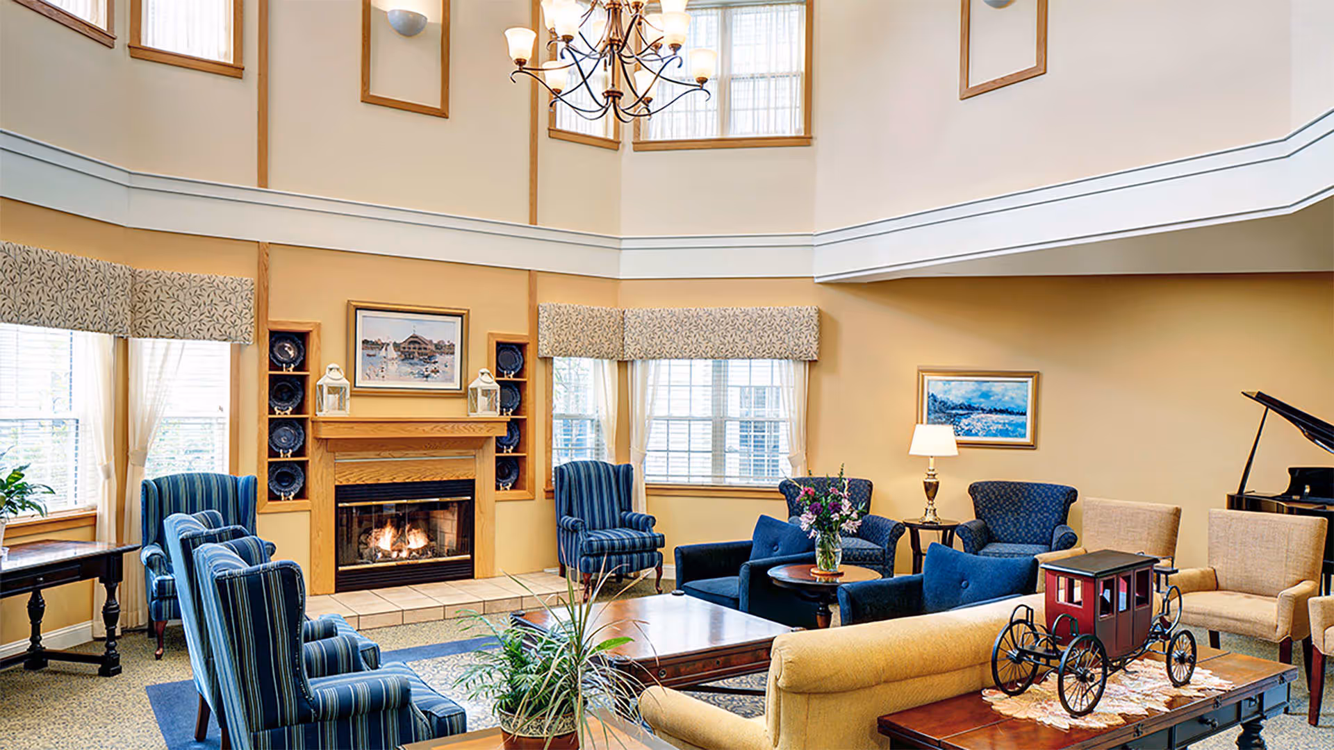 A cozy living room with a lit fireplace, surrounded by multiple upholstered chairs and sofas in blue and beige tones. The room features large windows with patterned valances, a chandelier hanging from a high ceiling, framed artwork on the walls, a coffee table with a vase of flowers, and a decorative model of a red carriage on a side table. A grand piano is visible in the background.