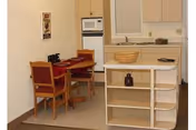 Small kitchen area with white cabinets, a microwave, and a countertop. In front of the kitchen is a small island with shelves and a decorative basket on top. To the left, there is a small dining table with two wooden chairs that have red cushions.