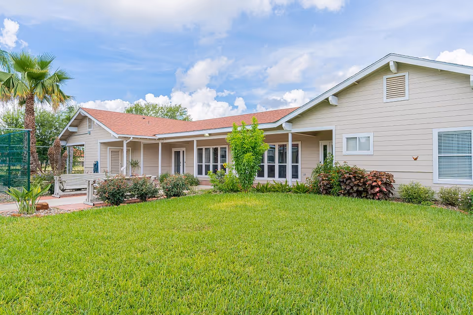 Exterior view of a single-story assisted living home with beige siding and a red roof, surrounded by green grass, shrubs, and palm trees under a partly cloudy sky.