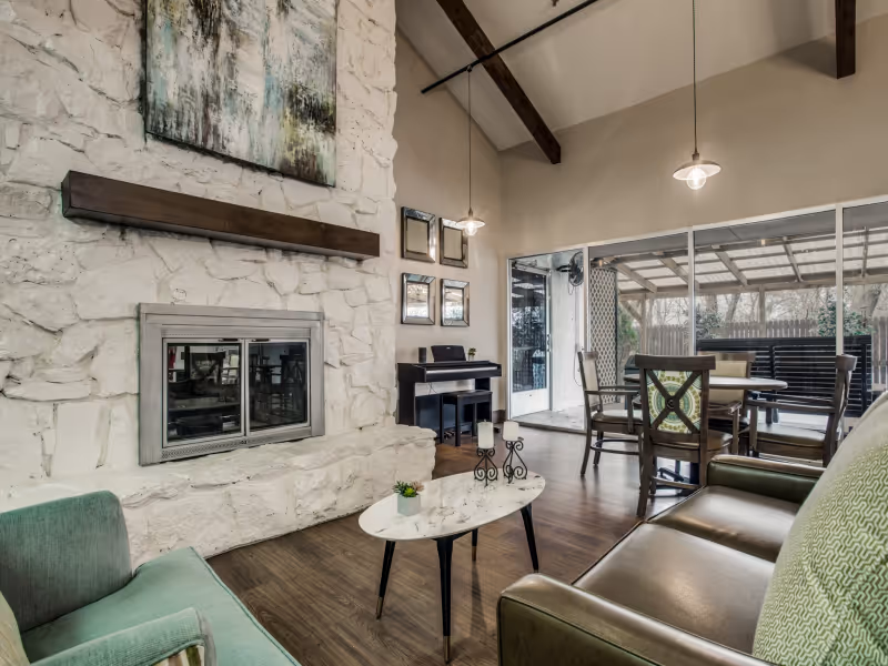 A cozy living room with a white stone fireplace, a wooden mantel, and a painting above it. The room features a green armchair, a brown leather sofa, and a small white marble coffee table with candles and a small plant. There is a piano against the wall with framed pictures above it. Large sliding glass doors open to an outdoor covered patio with a table and chairs. The room has wooden flooring and exposed wooden beams on the ceiling.