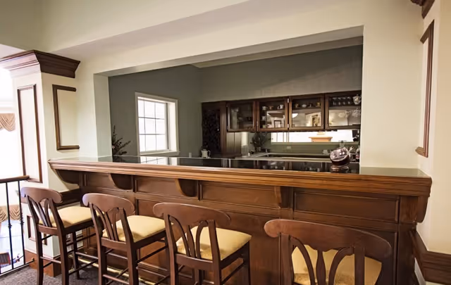 Interior view of a wooden bar counter with four high chairs featuring cushioned seats. Behind the bar is a kitchen area with cabinets, a window, and a countertop. The setting has a warm, classic design with wood paneling and neutral-colored walls.