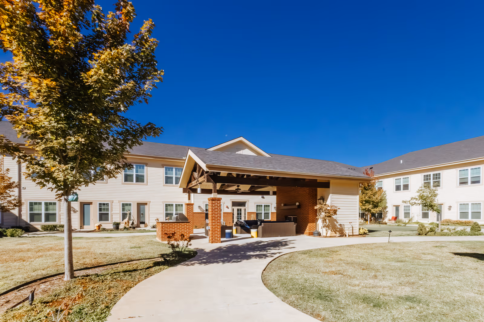 Outdoor courtyard area of The Harrison of Oklahoma City Assisted Living & Memory Care with a covered seating pavilion, surrounded by a two-story building, trees, and a clear blue sky.