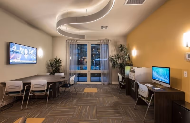 A modern interior room with a large table and several chairs on the left side, a wall-mounted TV displaying a news broadcast, and a desk with a computer and printer on the right side. The room has a patterned carpet with yellow accents, a large window with sheer curtains at the back, and decorative plants near the window. The ceiling features a unique curved light fixture.