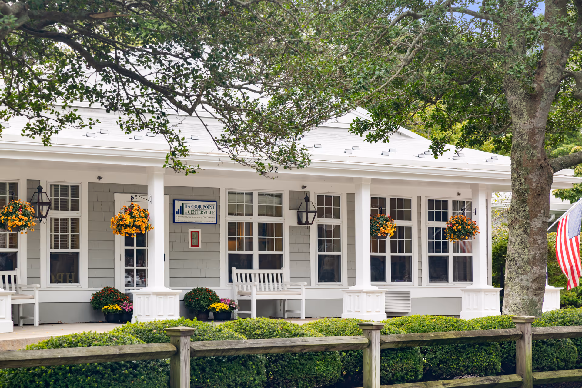 Front exterior view of Harbor Point at Centerville facility showing a white building with large windows, hanging flower baskets, white benches on the porch, a wooden fence, and an American flag on the right side. Trees and greenery surround the building.