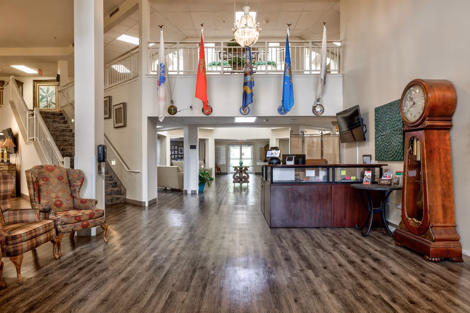 Interior view of a senior living facility lobby with wooden flooring, a reception desk on the right, a large grandfather clock, a wall-mounted TV, and several flags hanging from a balcony railing above. There are upholstered chairs on the left and a staircase leading to the upper floor.