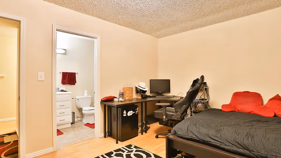 A small bedroom with a black bed and red pillows, a black office chair, a corner desk with a computer monitor, and a mini fridge underneath. The room has light-colored walls and wooden flooring. An open door reveals a bathroom with a toilet, white vanity, and red towels.