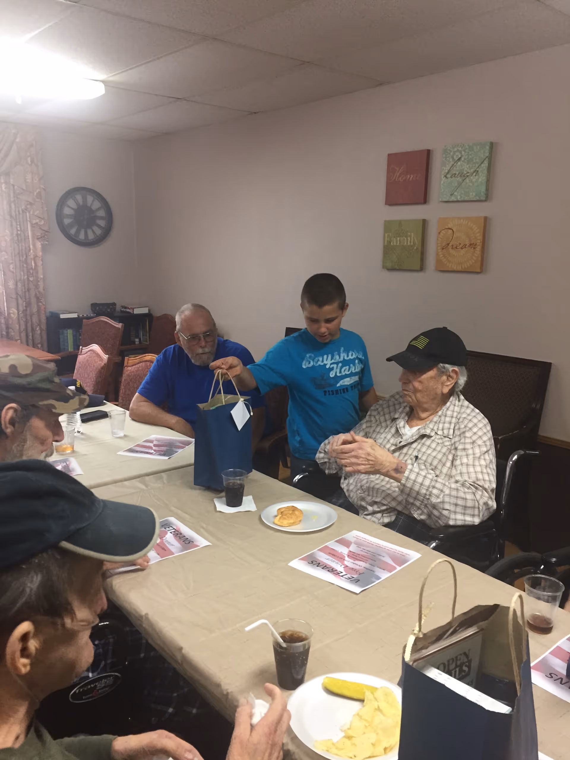 A group of elderly men and a young boy sitting around a table in a room. The boy is handing a gift bag to one of the elderly men who is seated in a wheelchair. The table has plates with food, drinks, and papers on it. The room has light-colored walls with four decorative wall plaques and a clock on the wall.