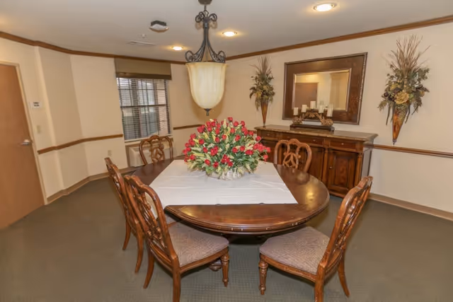 A dining room with a wooden oval table covered partially by a white tablecloth and a centerpiece of red flowers. Six wooden chairs with cushioned seats surround the table. On the wall behind the table is a large mirror flanked by two decorative wall arrangements. A hanging light fixture is above the table, and a window with blinds is visible on the left side.