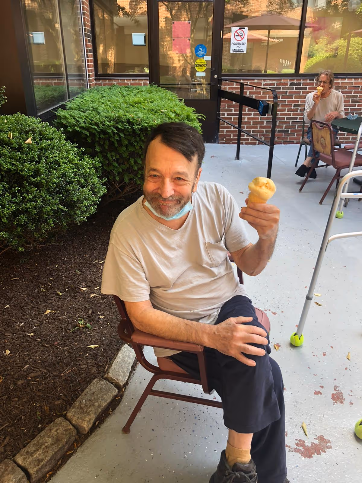 An elderly man sitting outdoors on a chair, smiling and holding an ice cream cone. In the background, another elderly person is seated at a table also eating ice cream. The setting appears to be a patio area with bushes, a brick wall, and glass doors.