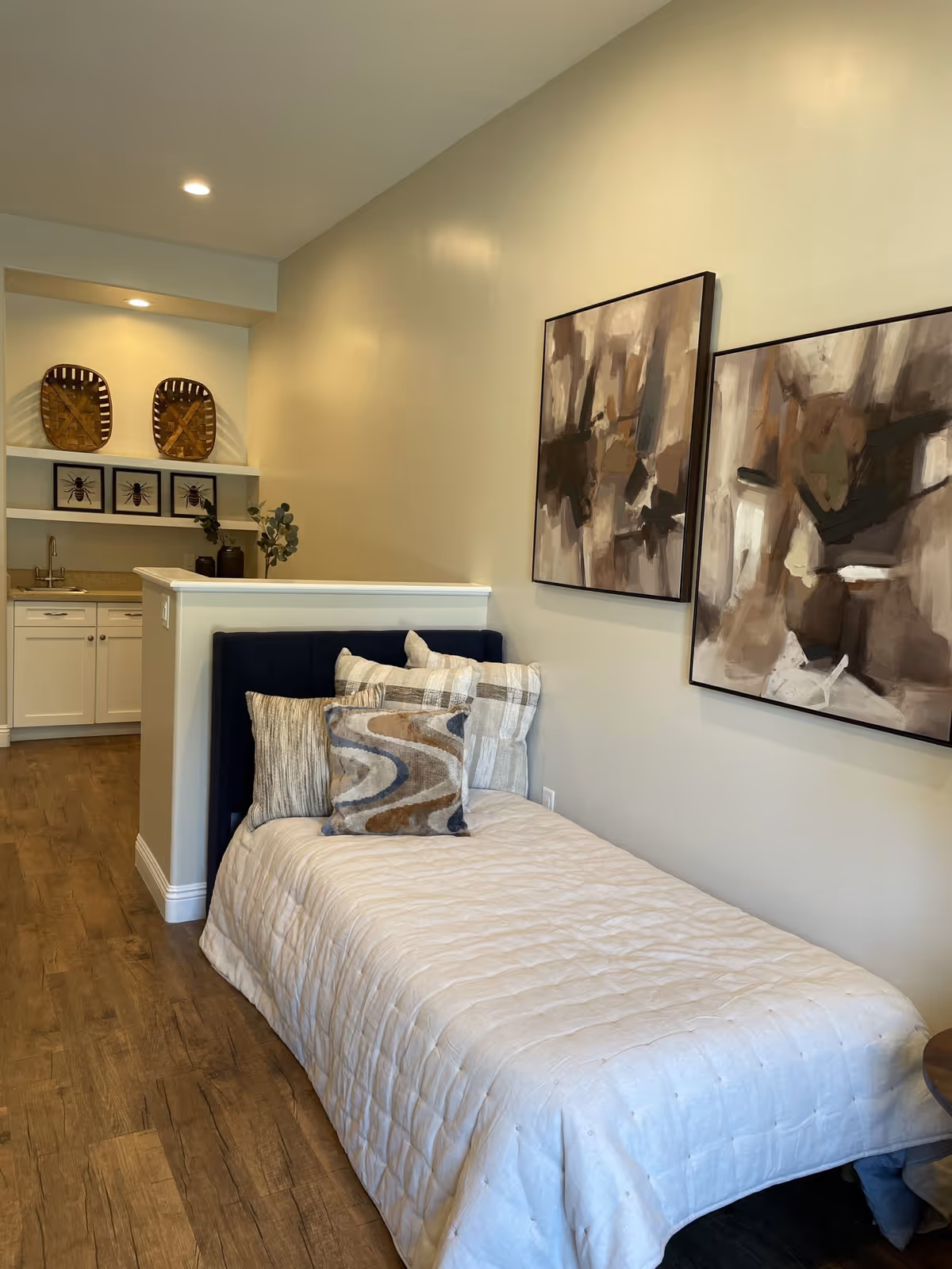 A small bedroom area with a single bed covered in a beige quilt and multiple decorative pillows. Two abstract paintings hang on the wall above the bed. In the background, there is a kitchenette with white cabinets, a sink, and decorative items including framed pictures and baskets on shelves. The floor is wooden, and the walls are painted a light beige color.