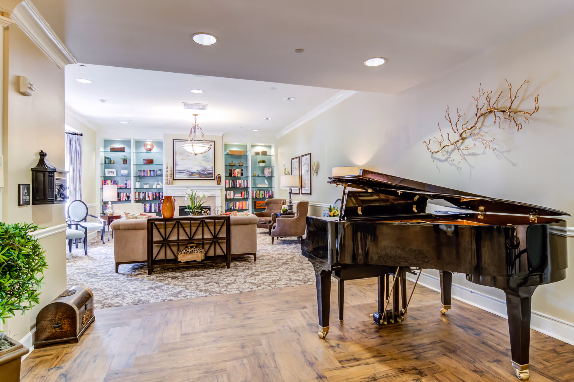 A bright and spacious living room area featuring a black grand piano on a wooden floor in the foreground. The background shows a cozy seating area with beige sofas and armchairs arranged around a fireplace with built-in bookshelves filled with books and decorative items. The room is decorated with plants, framed artwork, and a large wall sculpture resembling tree branches.