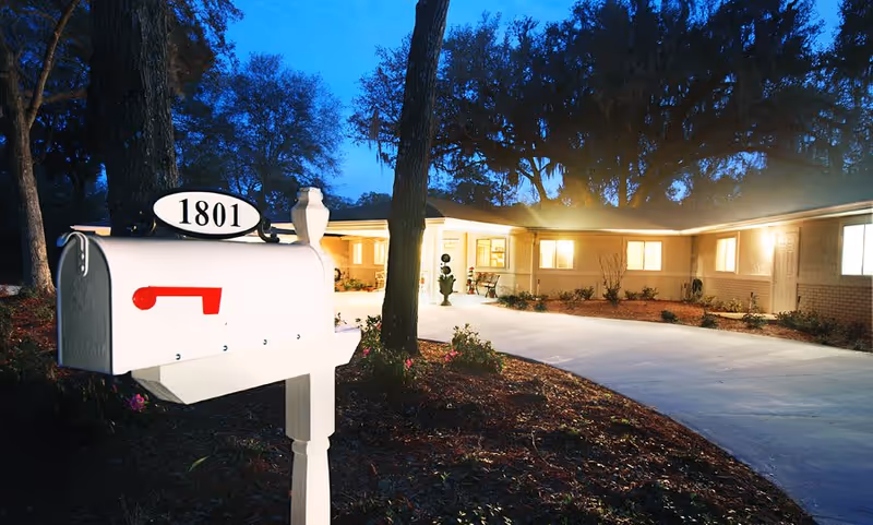 A white mailbox with the number 1801 in black on an oval sign is in the foreground. Behind it is a well-lit single-story building with multiple windows and a driveway leading up to it. The scene is set at dusk with trees and a dark blue sky in the background.
