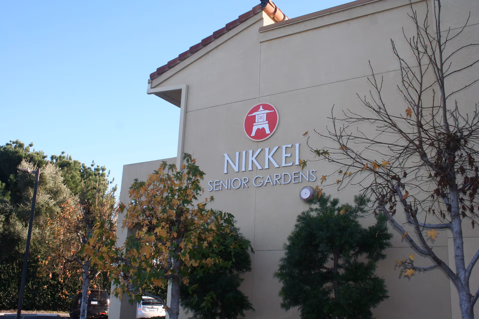 Exterior view of a building with the sign 'Nikkei Senior Gardens' mounted on a beige wall, surrounded by trees with some autumn leaves and a clear blue sky.