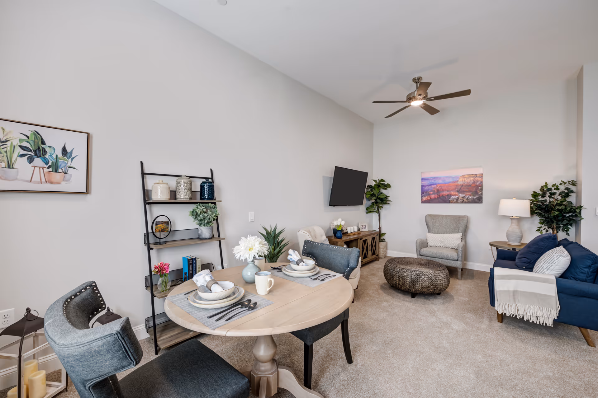 Bright furnished living room with a small round dining table, chairs, sofa, armchair, wall-mounted TV, shelving and decorative plants.