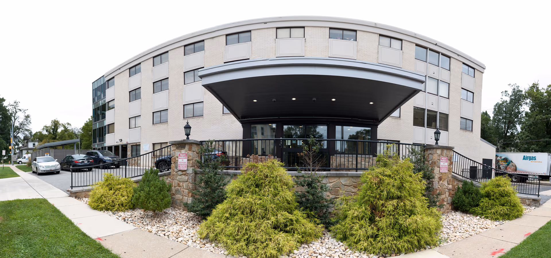 Front exterior view of a three-story senior living facility building with a covered entrance, stone pillars, landscaped bushes, and a parking area with several cars. The building has multiple windows and a light-colored facade.