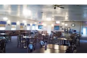 A spacious dining room with multiple round and rectangular tables and chairs arranged neatly. The room has large windows with blue valances, ceiling fans, and wall-mounted lights. There is a clock on the far wall and a serving area or counter in the background.