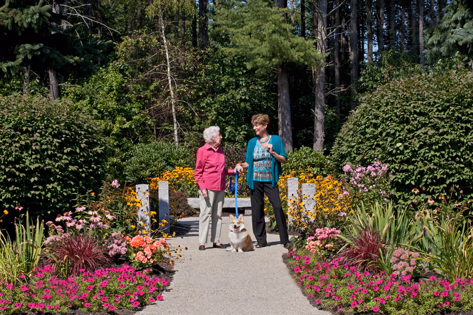 Two elderly women standing on a garden path surrounded by colorful flowers and greenery, with one woman holding a dog on a leash. Tall trees are visible in the background.