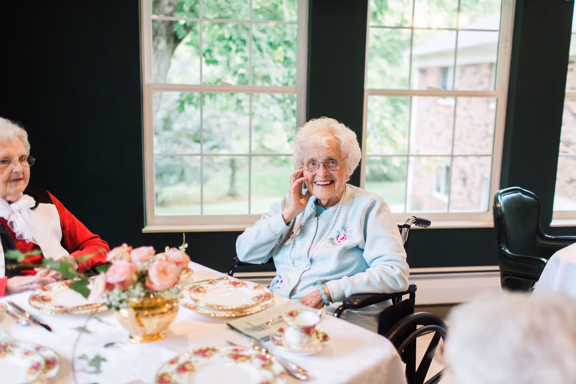 An elderly woman in a wheelchair smiling at a table set with floral china and a rose centerpiece in a bright dining room.