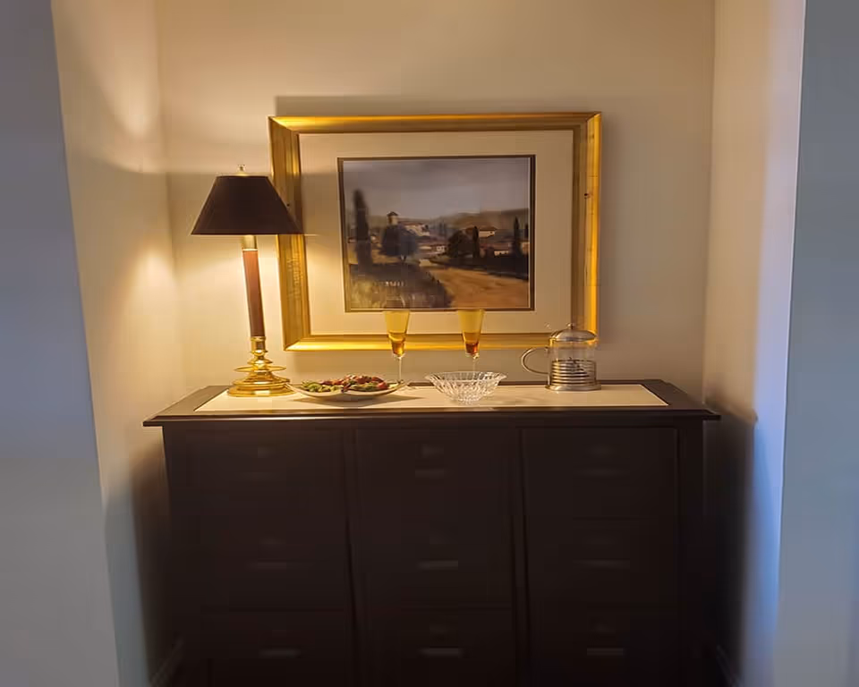 A dark wooden sideboard with multiple drawers is placed against a beige wall. On top of the sideboard, there is a table lamp with a dark shade and gold base, two amber-colored glasses, a small plate with food, a clear glass bowl, and a French press coffee maker. Above the sideboard hangs a framed landscape painting with a gold frame. The scene is softly lit by the lamp.