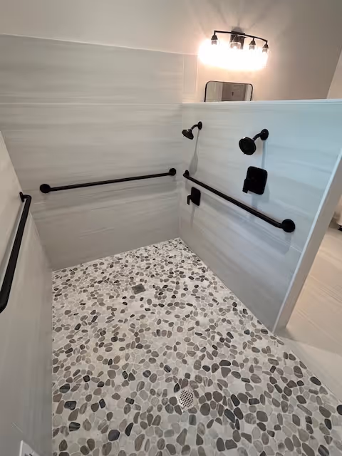 A modern walk-in shower with pebble stone flooring and light gray tiled walls. The shower features multiple black grab bars and a black showerhead and controls. Above the half wall is a light fixture with four bulbs and a small mirror.