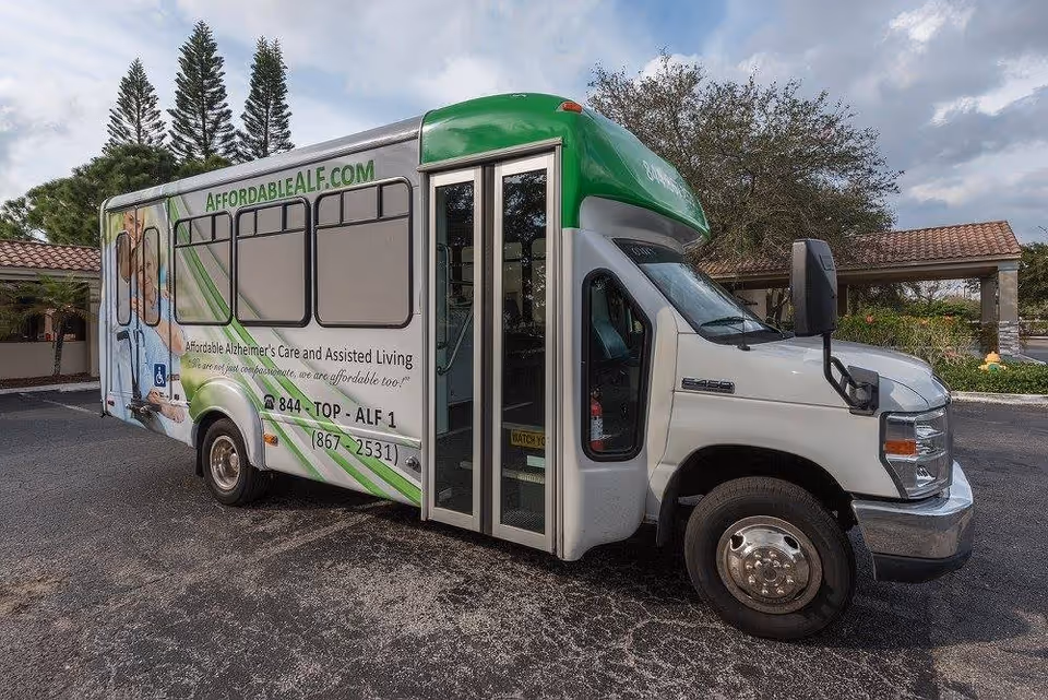 A white and green shuttle bus parked on an asphalt driveway in front of a building with a tiled roof and surrounded by trees. The bus has signage for AffordableALF.com, advertising Affordable Alzheimer's Care and Assisted Living with a phone number displayed.