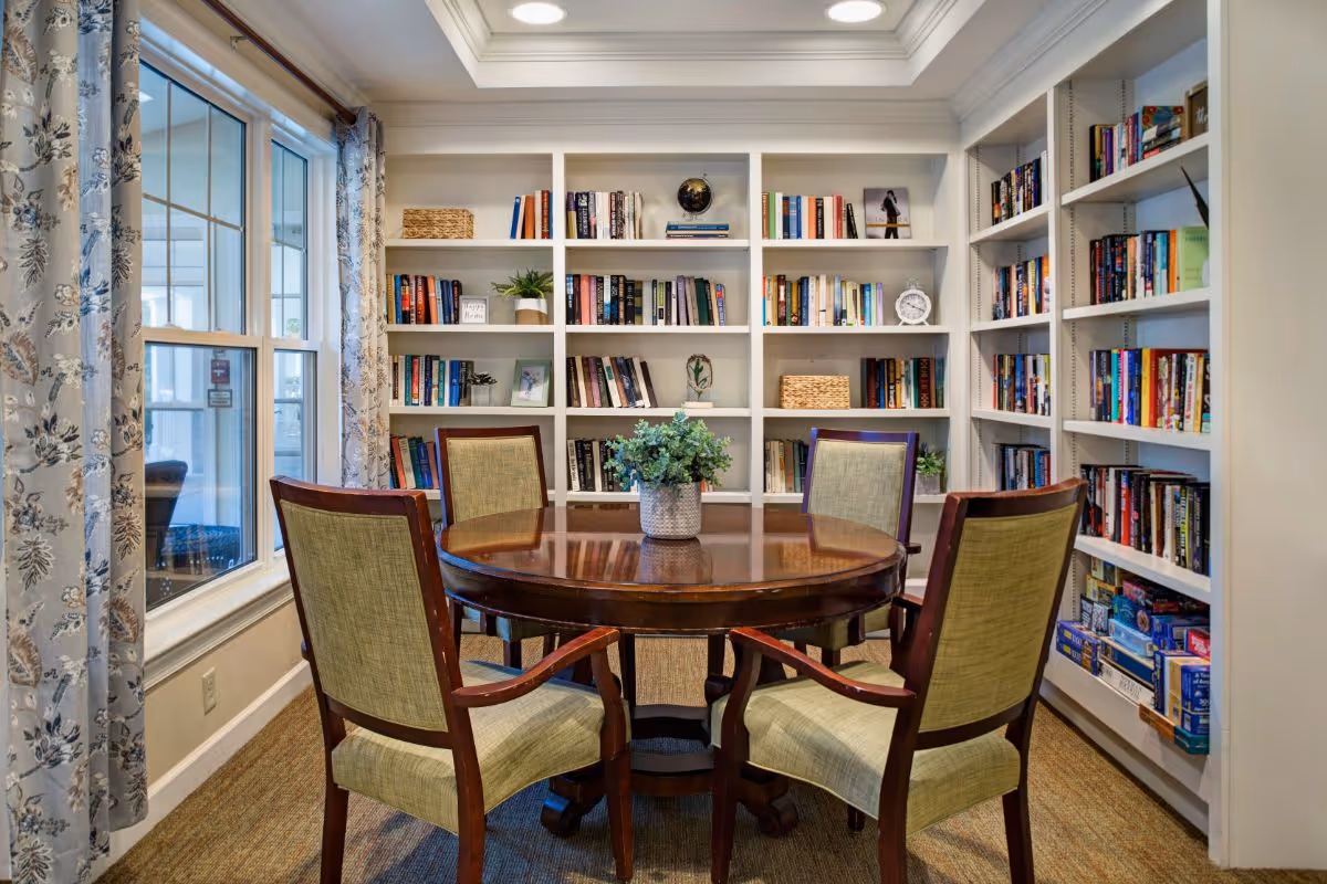 A cozy reading room with a round wooden table surrounded by four upholstered chairs. The walls are lined with white bookshelves filled with books, decorative items, and plants. A window with floral curtains lets in natural light.