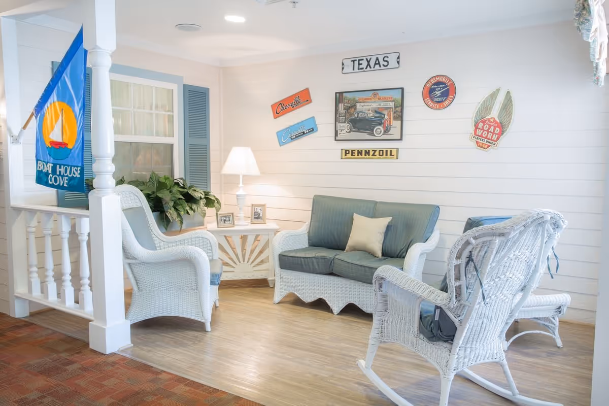 A cozy sitting area with white wicker furniture including a loveseat, two armchairs, and a rocking chair. The loveseat has blue cushions and a beige pillow. A small white side table with a lamp and framed photos is placed between the loveseat and one armchair. The walls are decorated with vintage signs and a framed picture, and a blue flag with a yellow sailboat and the words 'Boat House Cove' hangs on a white column. The floor is wood with a carpeted area nearby.