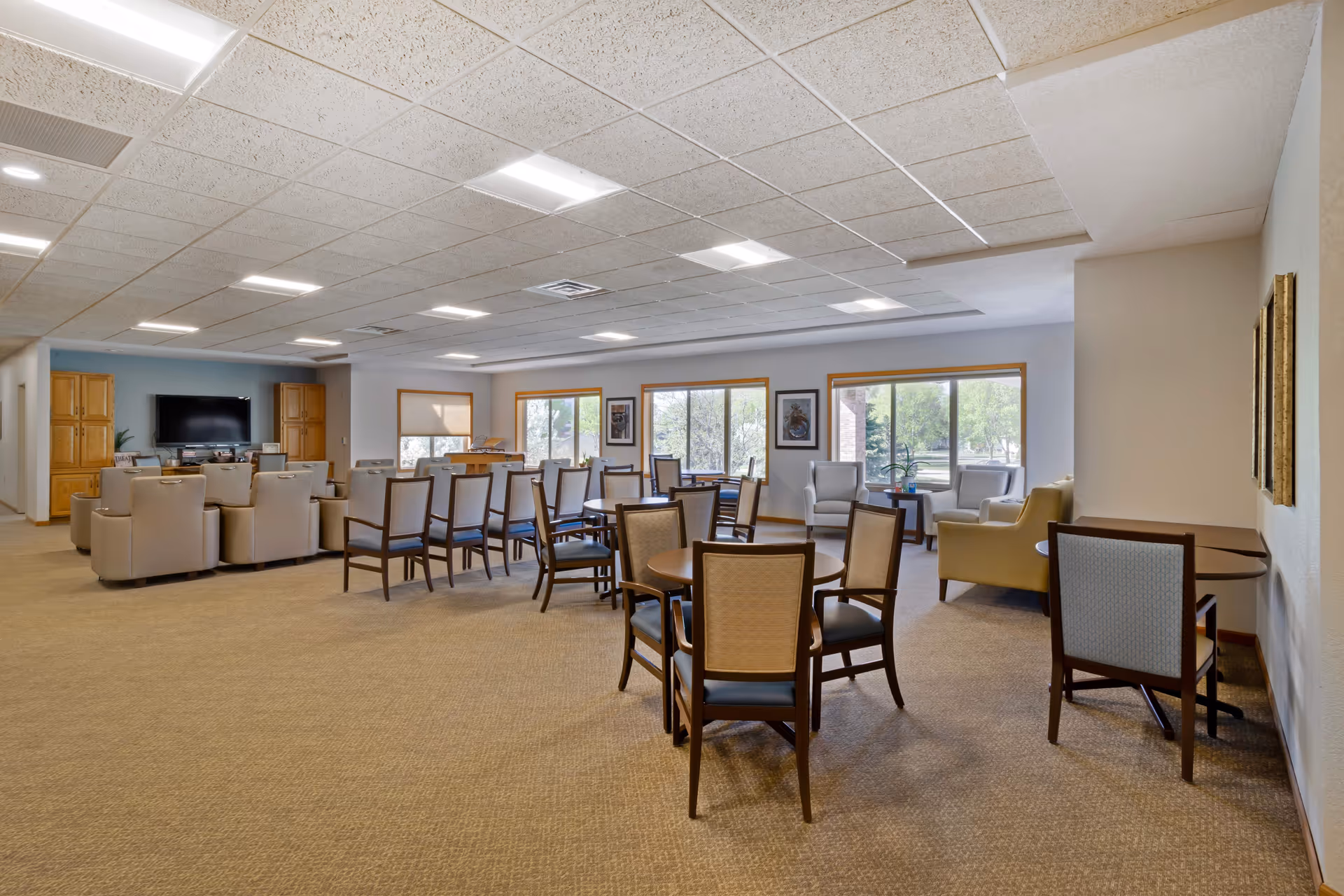 A spacious senior living common area with multiple seating arrangements including round tables with chairs, armchairs, and a row of recliners facing a large flat-screen TV mounted on a light blue accent wall. The room has large windows letting in natural light and framed artwork on the walls.