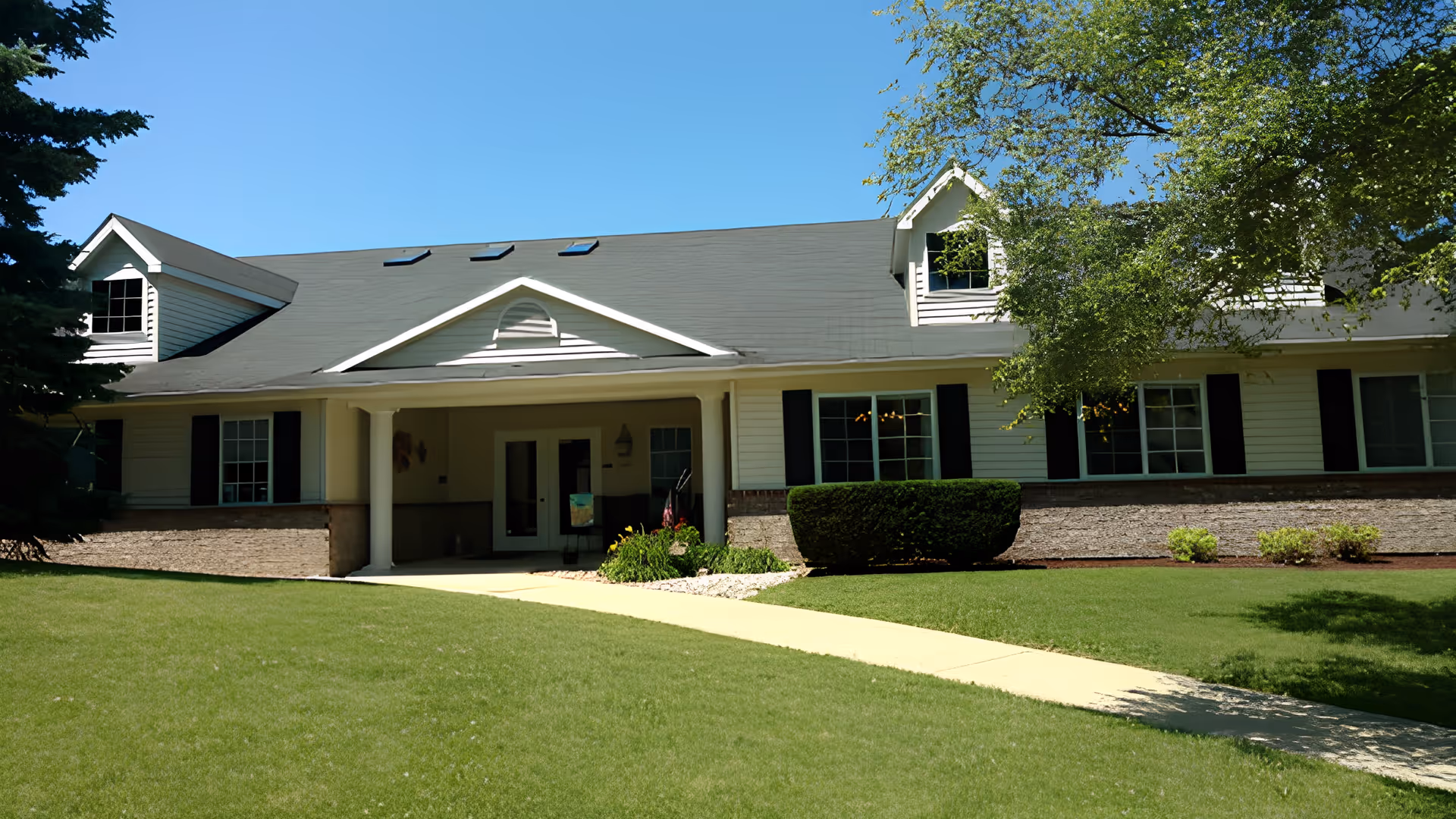 Front exterior view of a single-story senior living facility building with a gray roof, white siding, black window shutters, and a covered entrance. The building is surrounded by green grass, bushes, and trees under a clear blue sky.