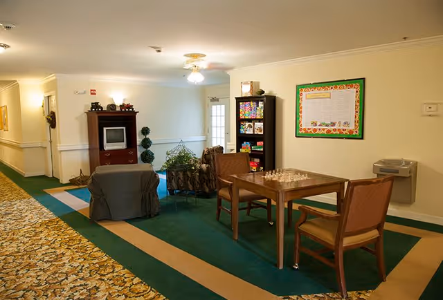 A cozy common area in a senior living facility featuring a small wooden table with a chessboard and two chairs, a bookshelf stocked with games and books, a vintage TV on a wooden cabinet, two armchairs, a water fountain on the wall, and a bulletin board. The floor is carpeted with green and beige patterns, and the walls are painted light yellow.