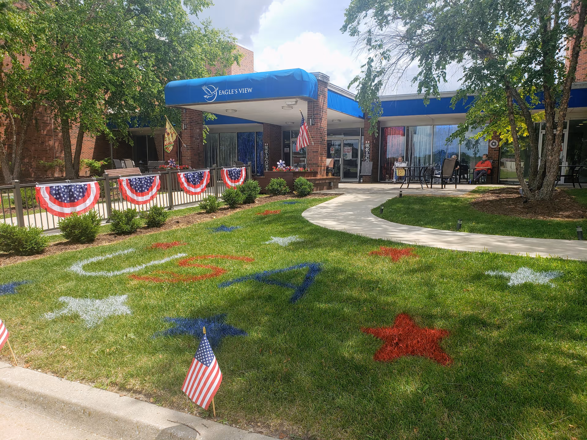 Front entrance of Eagle's View Supportive Living and Memory Care facility with a blue awning displaying the facility's name. The lawn in front is decorated with red, white, and blue stars and the letters 'USA' painted on the grass. American flags and patriotic bunting adorn the railing and lawn. Several people are seated on chairs near the entrance under the awning. Trees and a clear sky are visible in the background.