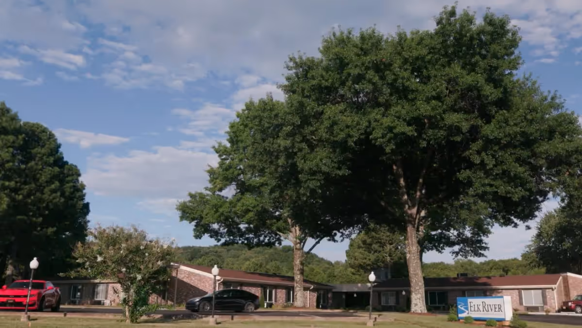 Exterior view of Elk River Health and Nursing Center of Fayetteville showing a single-story brick building with a brown roof, surrounded by large green trees and a partly cloudy sky. There are a few cars parked in front and a sign with the facility's name near the entrance.