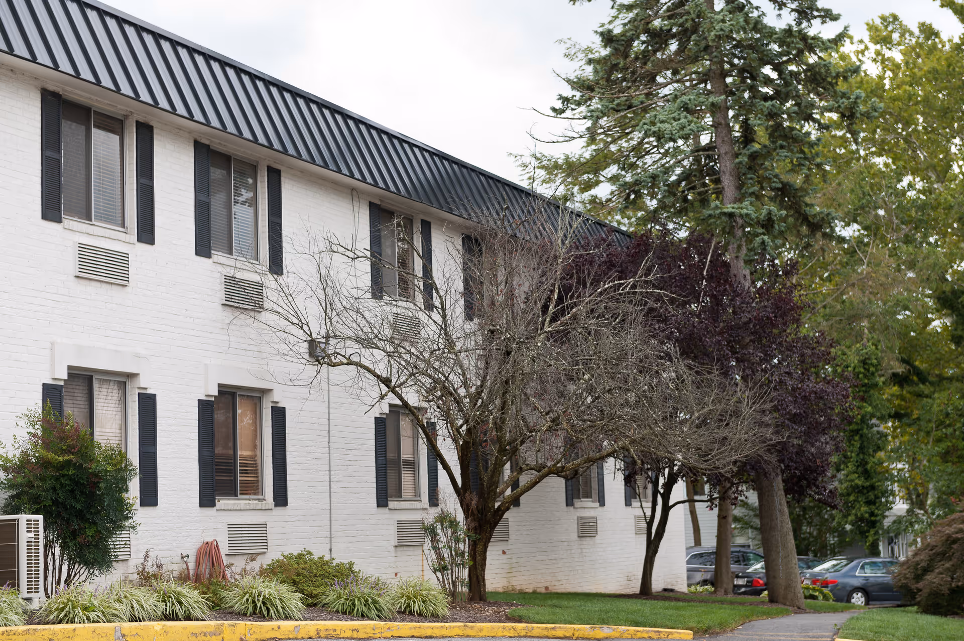 Exterior view of a two-story white brick building with black shutters and a black metal roof. There are several windows, some trees with sparse leaves, bushes, and a small parking area with cars visible in the background.