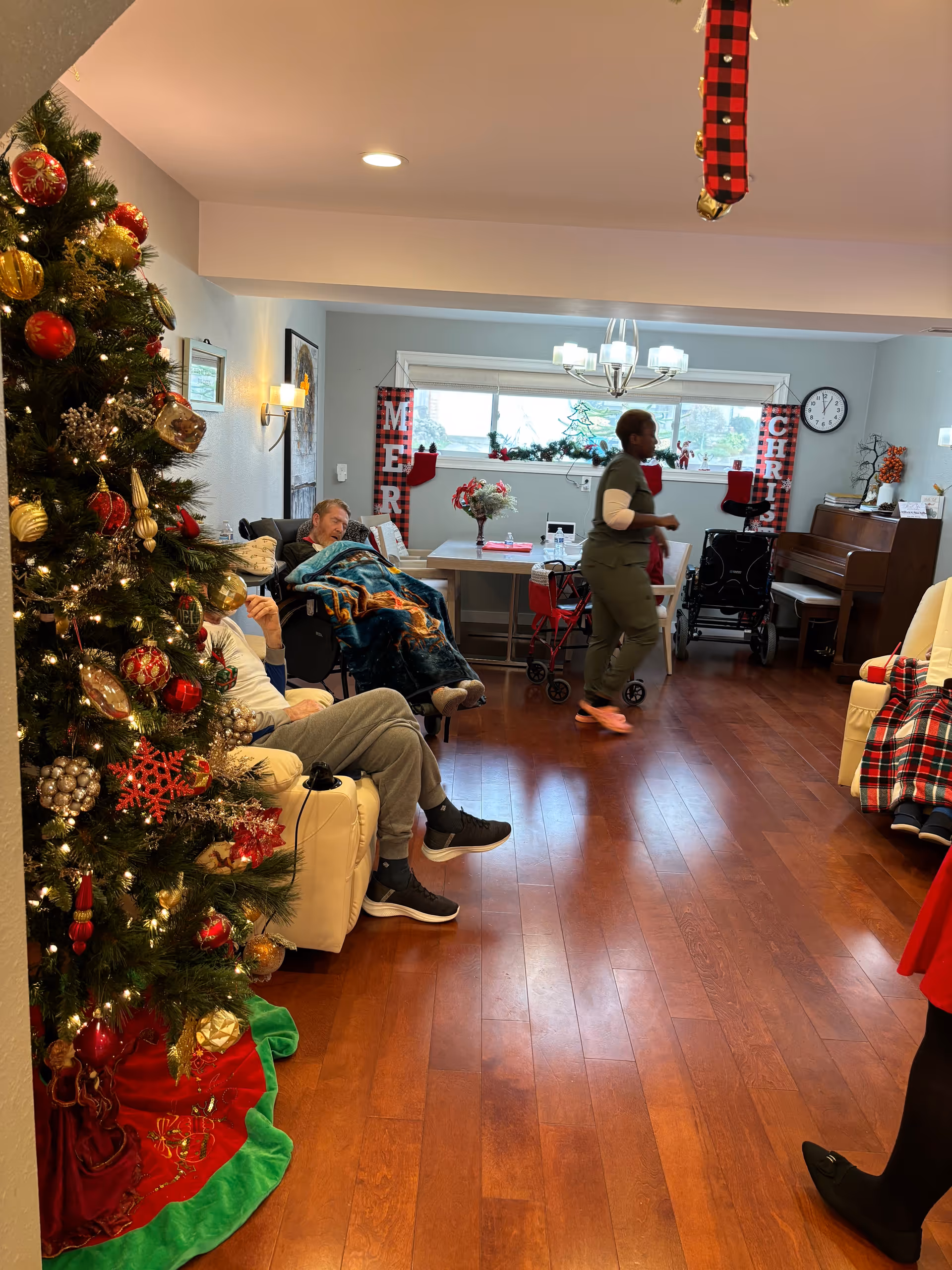 A cozy living room decorated for Christmas with a large decorated Christmas tree on the left. Two elderly people are seated on cream-colored recliners, one covered with a blanket. A caregiver in green scrubs walks across the room. The room has wooden floors, a piano in the back right corner, and a dining table with Christmas decorations near a window with holiday banners spelling 'MERRY CHRISTMAS'.