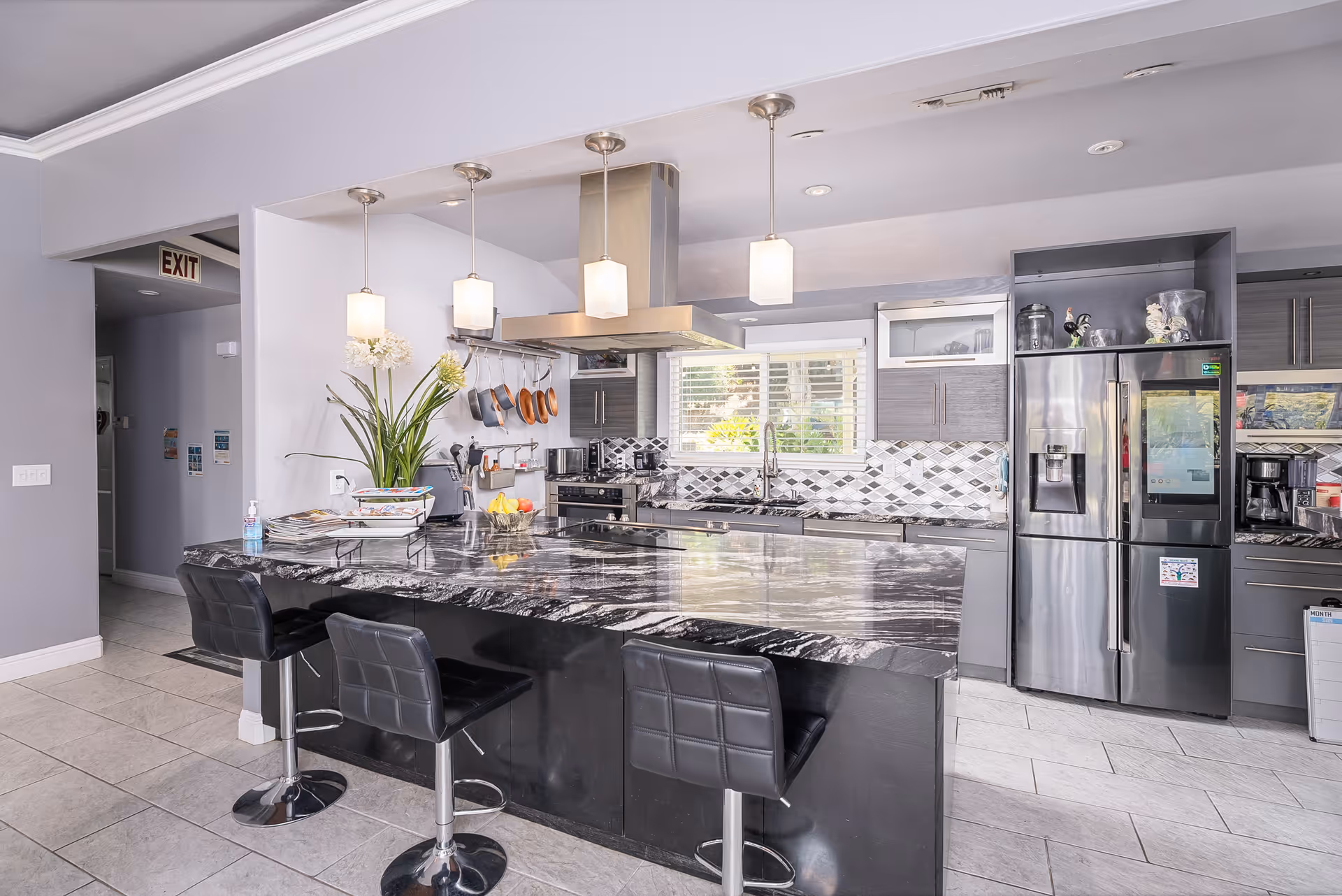Modern kitchen with a large black and gray marble island countertop featuring three black bar stools. The kitchen has gray cabinets, a stainless steel refrigerator with a digital screen, a stove with a stainless steel hood, and a window above the sink. Pendant lights hang above the island, and there are various kitchen utensils and plants on the counter.