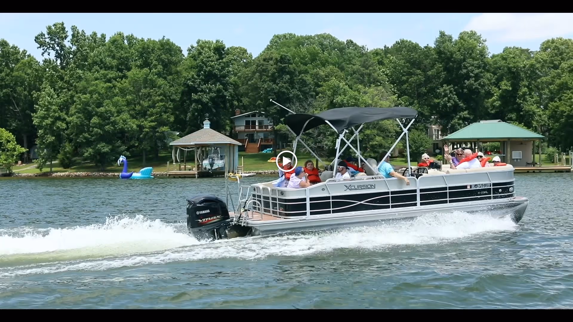 A group of people enjoying a ride on a pontoon boat named Xcursion on a lake, with green trees and lakeside houses in the background.
