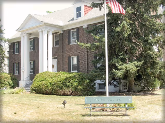 Two-story brick building with white columns at the entrance, an American flag on a pole, and a bench on the front lawn.
