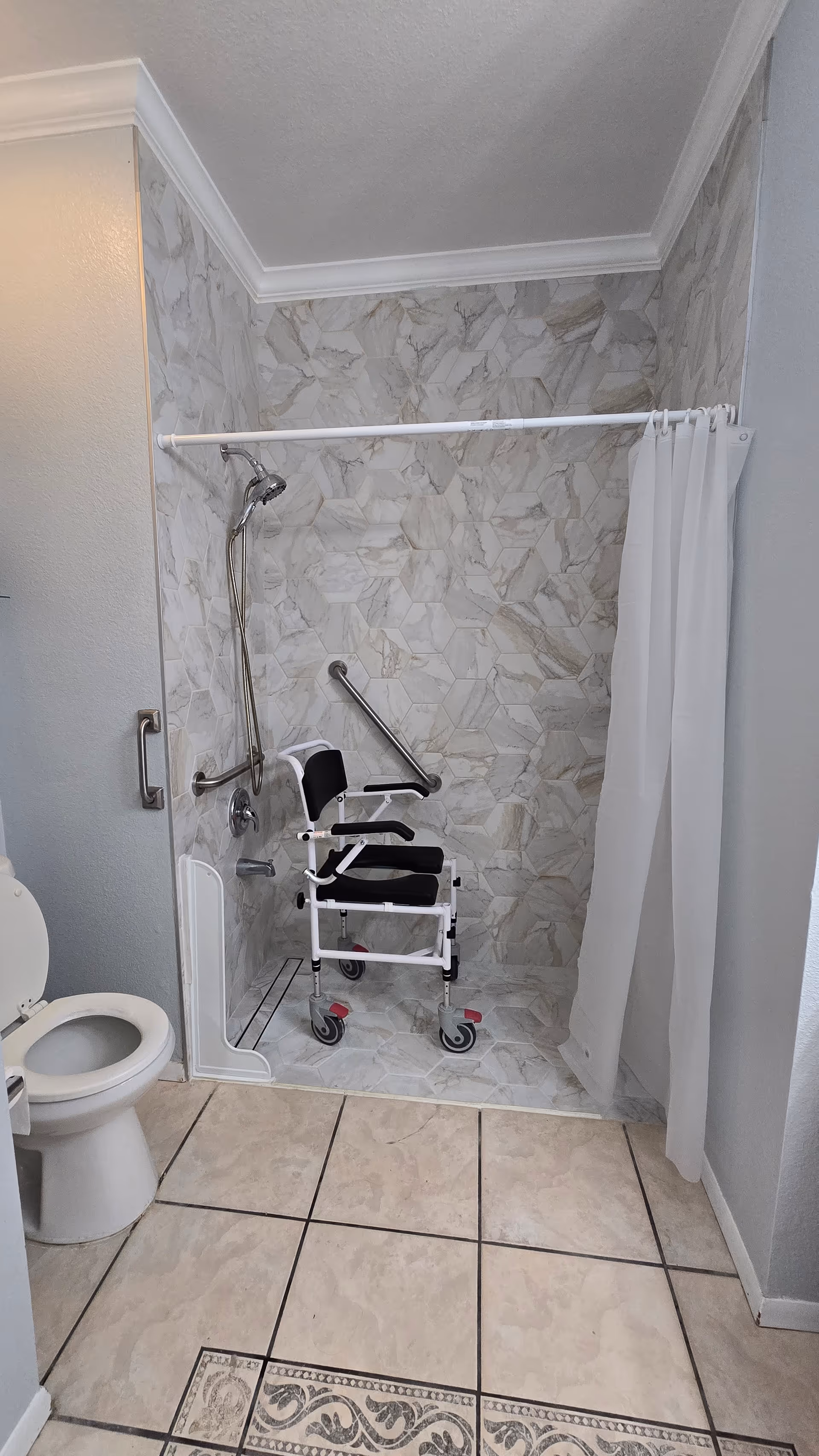 A bathroom with a tiled floor and a walk-in shower area featuring marble-patterned tiles. Inside the shower is a white and black shower chair with wheels and armrests. There are two metal grab bars mounted on the shower walls for support. A white shower curtain is partially drawn to the side. A white toilet is visible to the left of the shower.