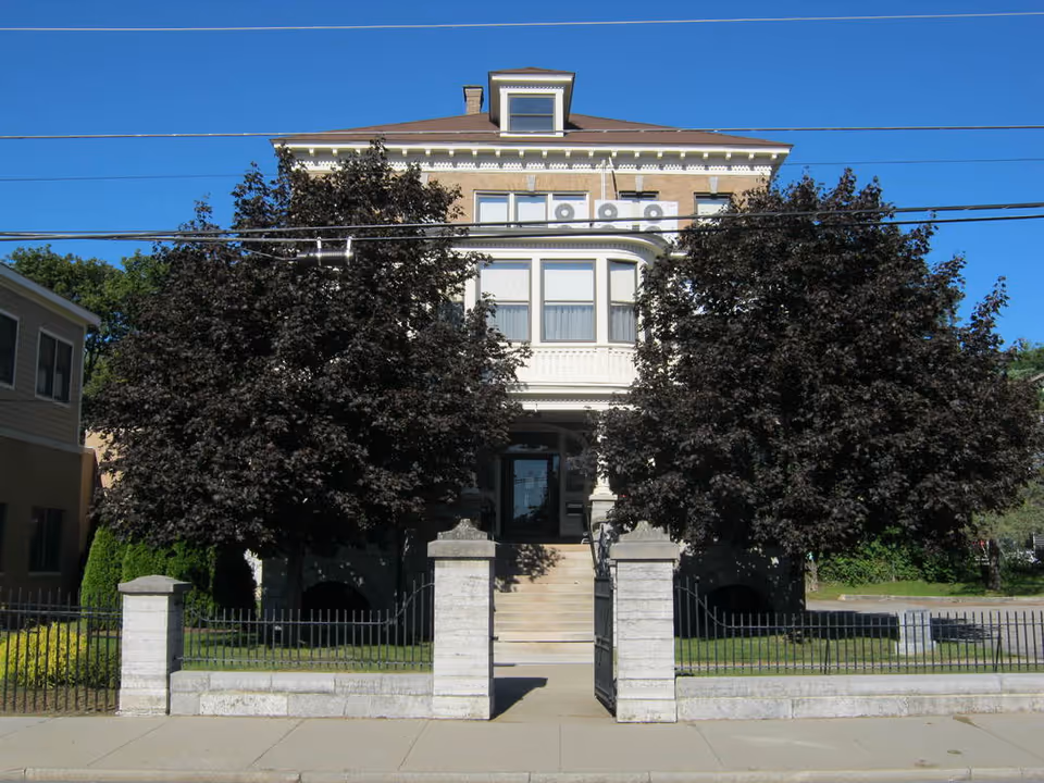 Front exterior view of a multi-story brick building with a small staircase leading to the entrance, flanked by two large leafy trees and a black metal fence with stone pillars in front.