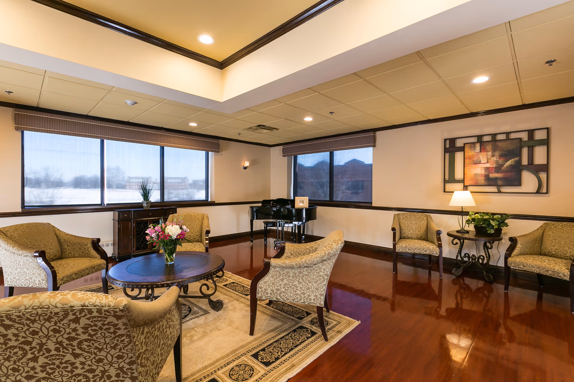 A well-lit sitting area in a nursing and rehabilitation center featuring patterned upholstered armchairs arranged around a round wooden coffee table with a vase of flowers. The room has large windows with blinds, a polished wooden floor, a decorative rug, a side table with a lamp and plant, a framed abstract painting on the wall, and a black piano in the corner.