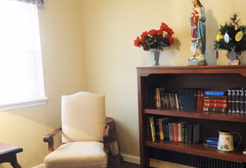 A cozy corner of a room featuring a cushioned armchair next to a wooden bookshelf filled with books. On top of the bookshelf, there is a vase with red flowers, a statue, and a small decorated tree. A window with blinds is visible on the left side, allowing natural light to brighten the space.