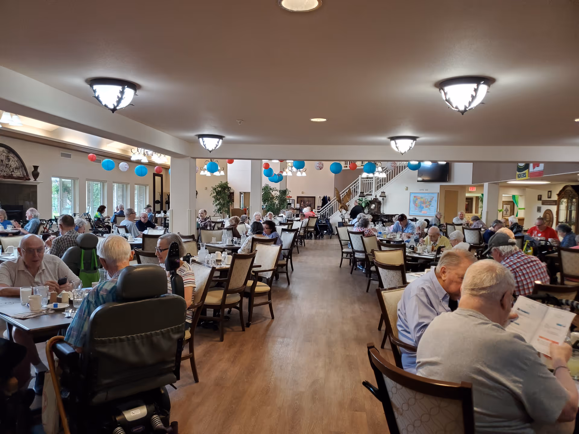 A large dining room in a senior living facility with many residents seated at tables and colorful hanging decorations.