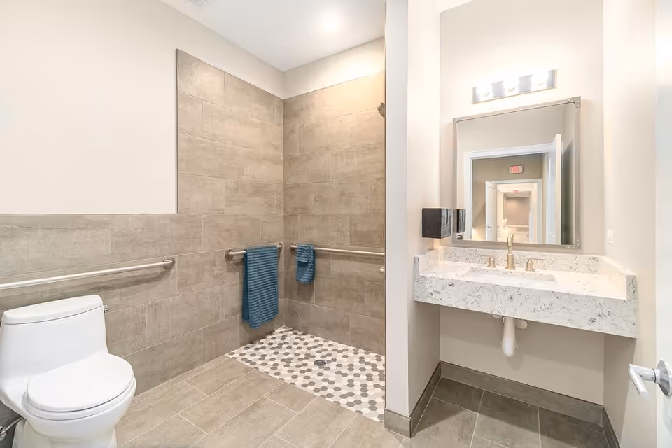 A modern, accessible bathroom featuring a white toilet, a walk-in shower with gray and white hexagonal floor tiles, beige wall tiles, and stainless steel grab bars with blue towels hanging on them. There is a white marble countertop with a sink and a large mirror above it, reflecting a hallway with multiple doors.