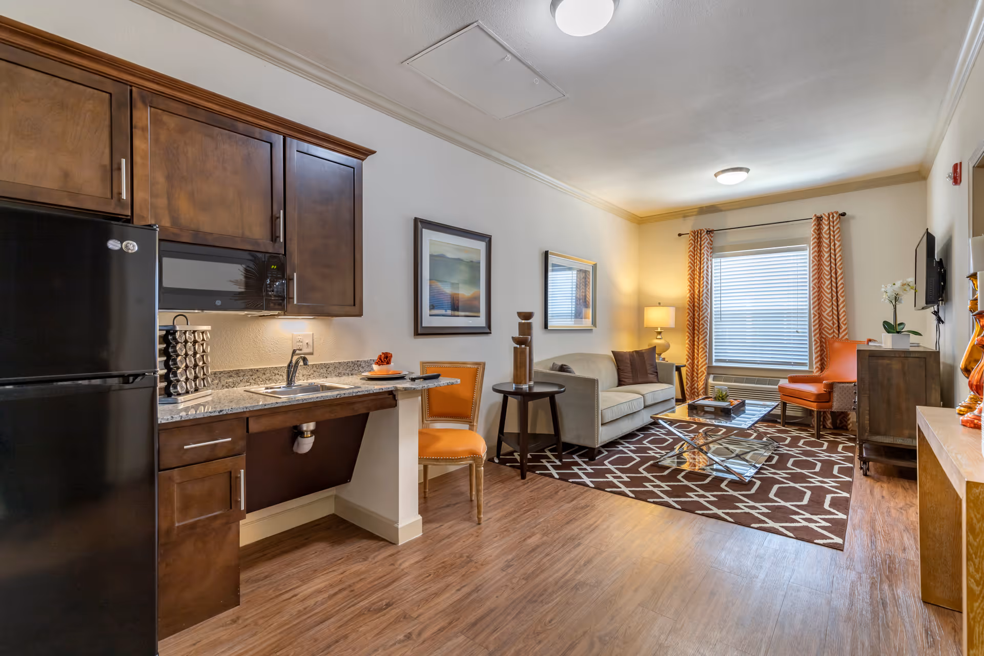 A cozy living space featuring a compact kitchen area with dark wood cabinets, a black refrigerator, and a granite countertop with a sink. Adjacent to the kitchen is a small dining area with an orange chair. The living room includes a beige sofa, a glass coffee table on a patterned rug, an orange armchair, a side table with a lamp, and a window with orange patterned curtains allowing natural light to fill the room.