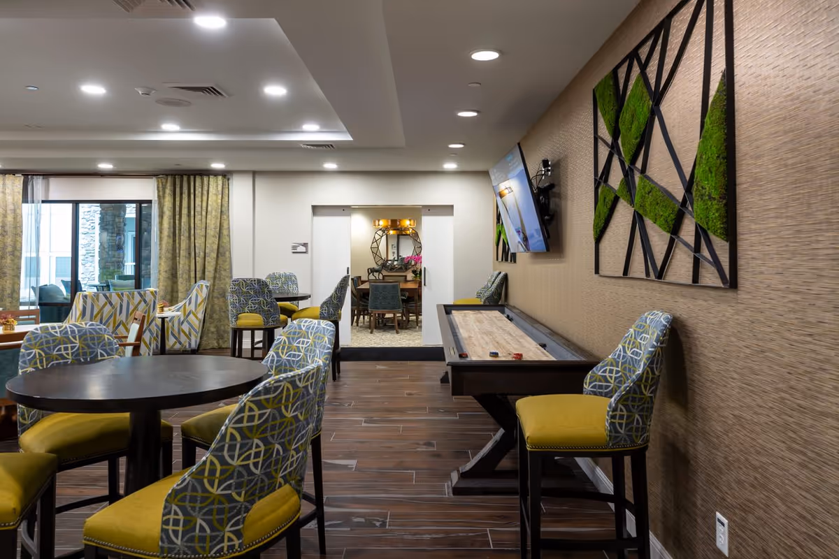 A modern common area with patterned chairs around dark wooden tables, a shuffleboard table along the right wall, and a flat-screen TV mounted above it. The room has wood flooring, recessed ceiling lights, and decorative green and black wall art. In the background, there is a doorway leading to another room with additional seating and a round mirror on the wall.