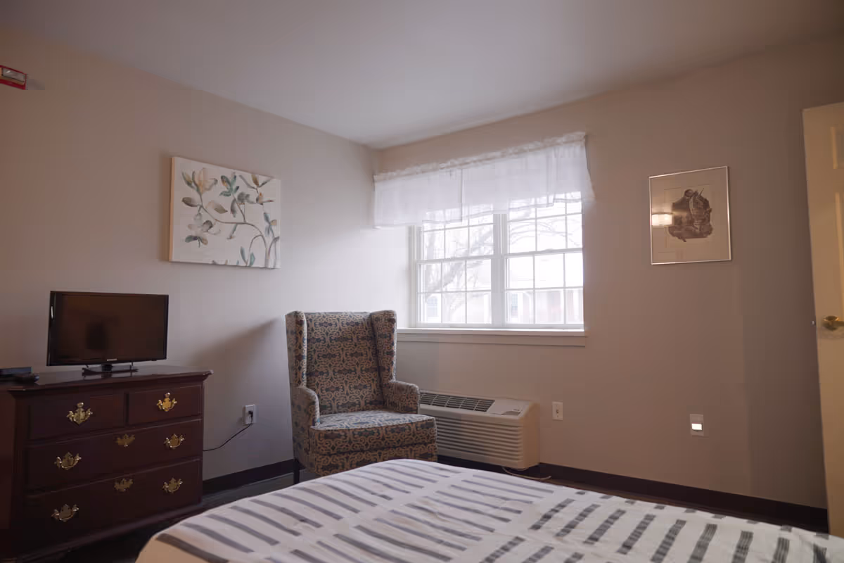 A bedroom with a bed in the foreground covered with a striped blanket, a patterned armchair near a window with sheer white curtains, a wooden dresser with a small flat-screen TV on top, and two framed pictures on the walls.